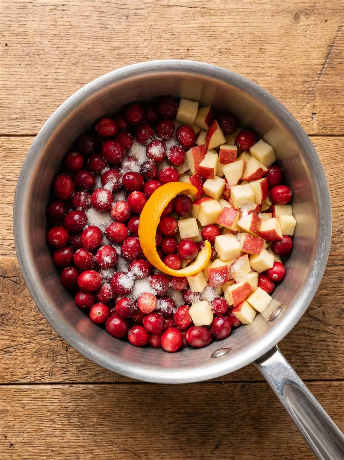 Fresh cranberries, diced apple, orange peel, and sugar in a saucepan before cooking.