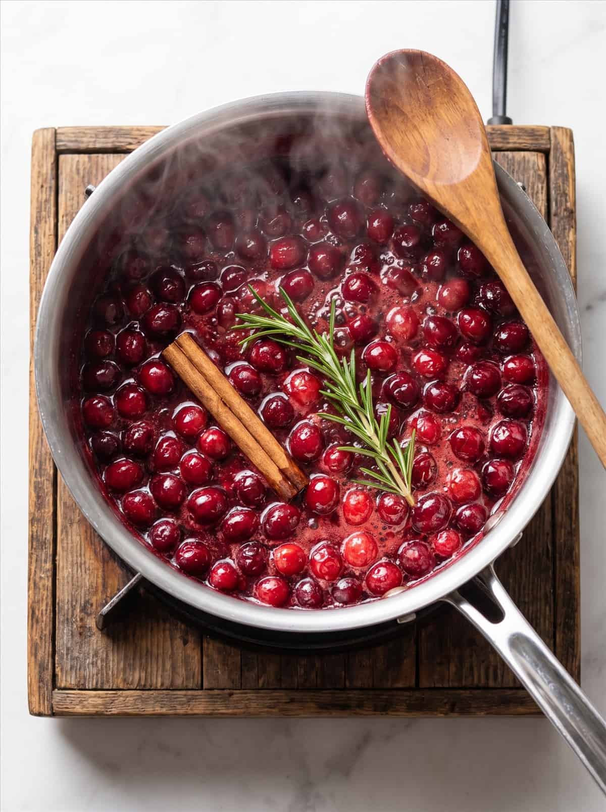 Cranberries bursting and simmering into a thick red sauce with cinnamon and rosemary in a saucepan.