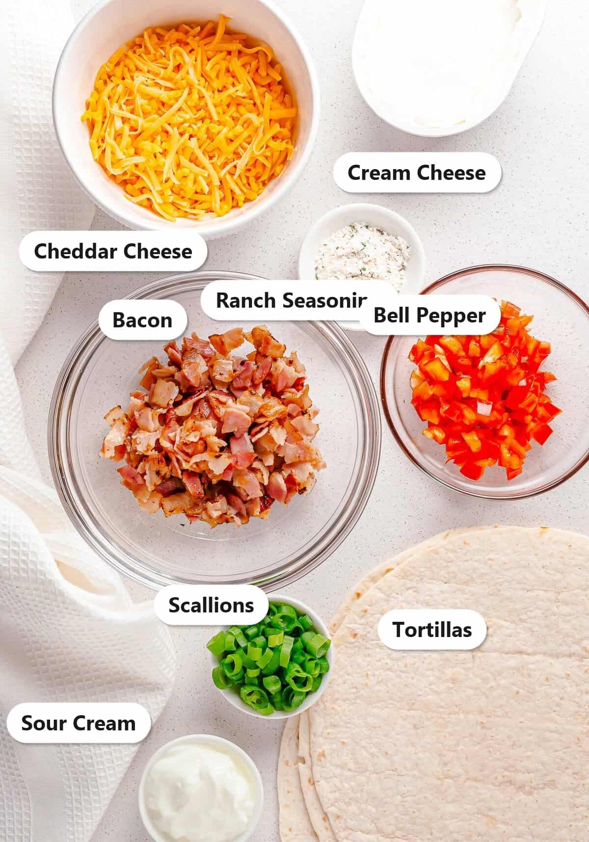 Labeled ingredients for cheddar bacon ranch cream cheese pinwheels including cream cheese, cheddar, bacon, bell pepper, scallions, sour cream, ranch seasoning, and tortillas.