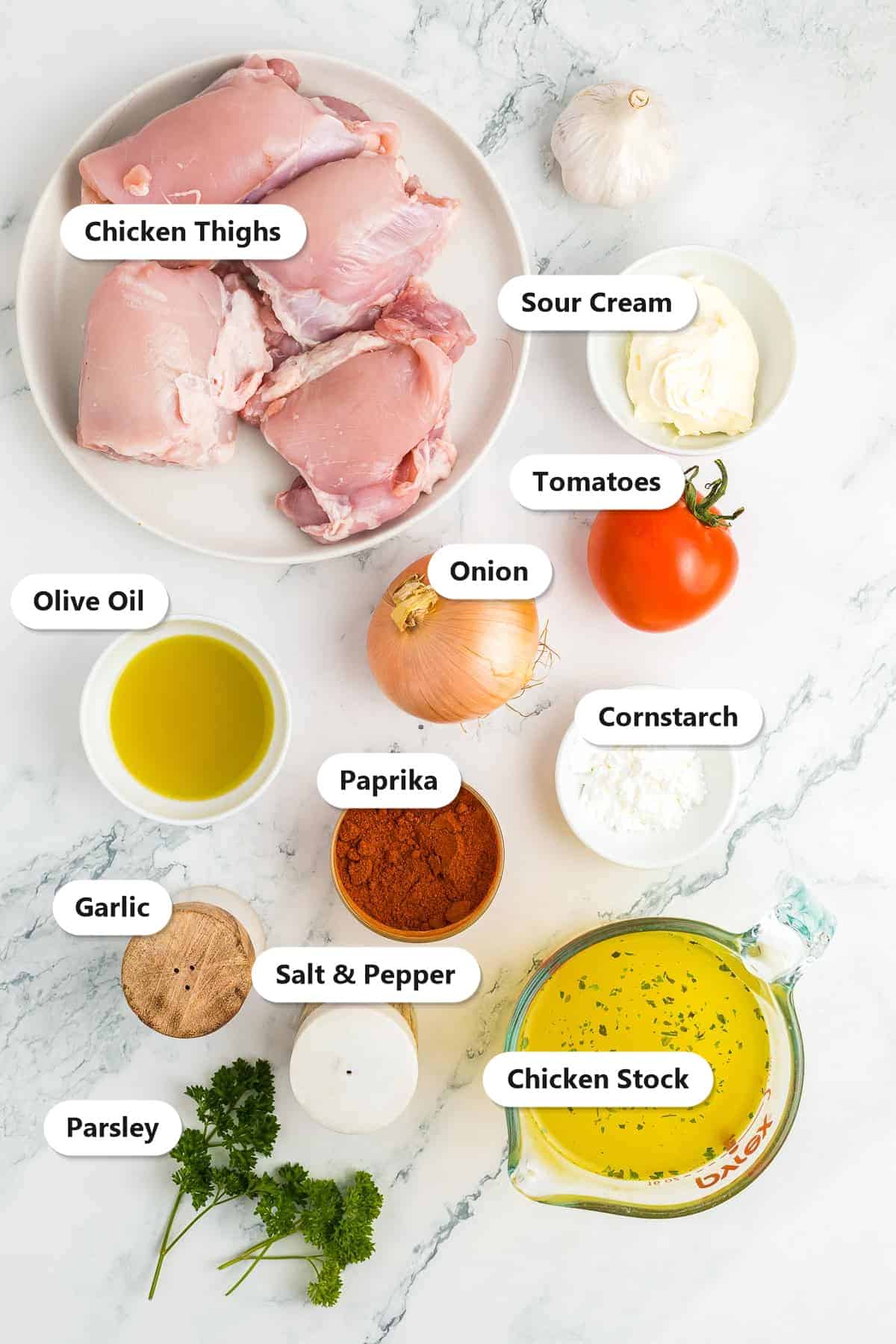 Labeled ingredients for creamy paprika chicken thighs including chicken thighs, sour cream, tomatoes, onion, olive oil, paprika, cornstarch, garlic, salt and pepper, chicken stock, and parsley.
