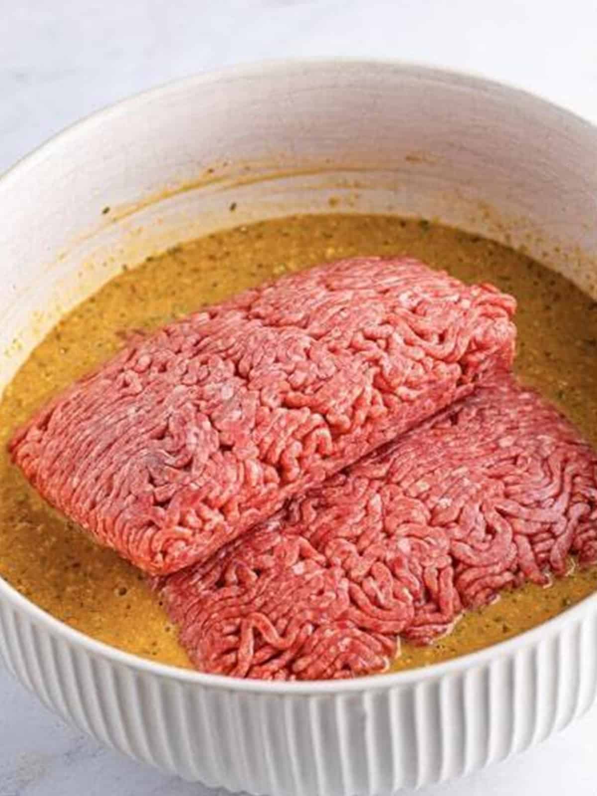 Lean ground beef added to the meatloaf base mixture in a white bowl.