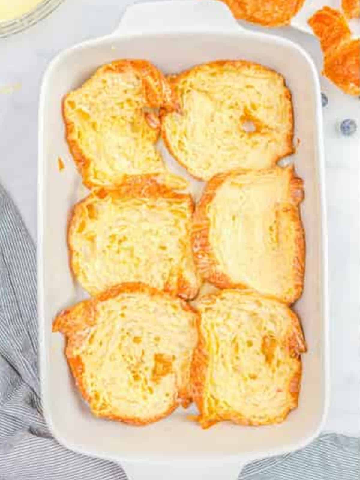 Dipped croissant halves arranged in a white baking dish for french toast.