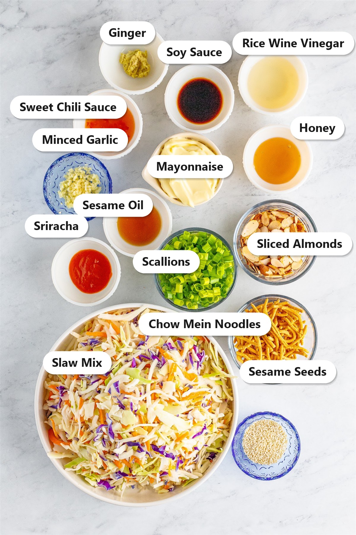 Labeled ingredients for crunchy Asian slaw including slaw mix, mayonnaise, rice wine vinegar, sweet chili sauce, honey, soy sauce, minced garlic, sriracha, ginger, sesame oil, scallions, sliced almonds, chow mein noodles, and sesame seeds.