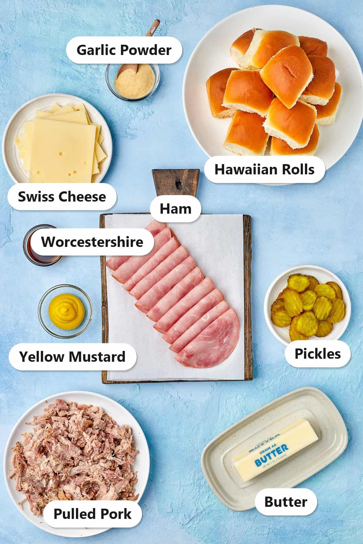 Labeled ingredients for Cuban sliders including Hawaiian rolls, Swiss cheese, ham, pulled pork, pickles, butter, mustard, Worcestershire, and garlic powder.