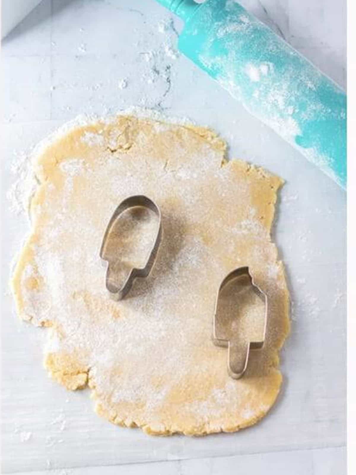 Rolled sugar cookie dough being cut into popsicle shapes with cookie cutters.