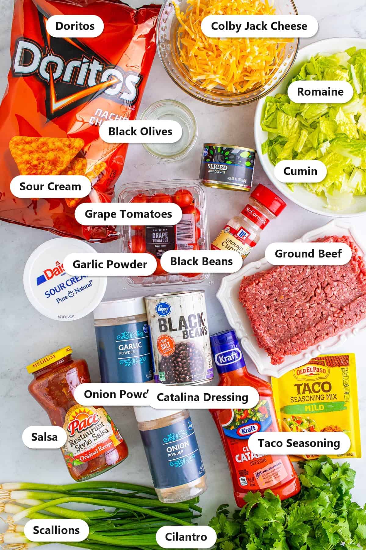Labeled ingredients for Doritos Taco Salad including ground beef, Doritos, cheese, black beans, tomatoes, olives, Catalina dressing, and taco seasoning.