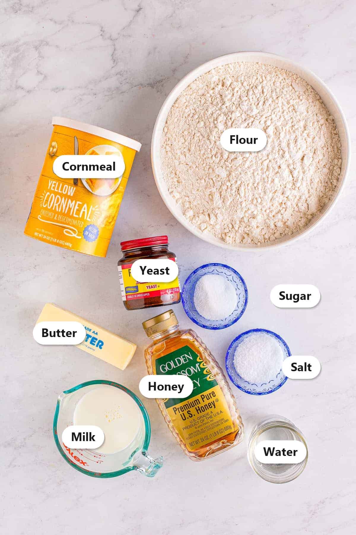 Labeled ingredients to make English muffins including flour, yeast, sugar, salt, milk, water, honey, butter, and cornmeal.
