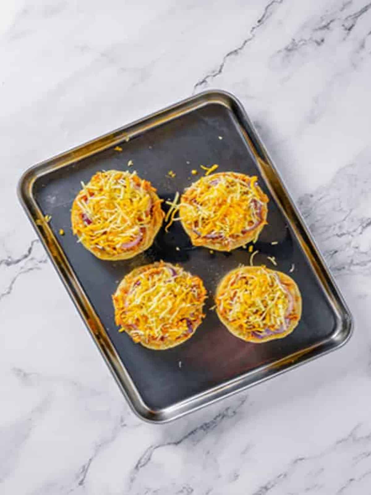 English muffin halves topped with BBQ chicken, red onion, and shredded cheese on a baking sheet.
