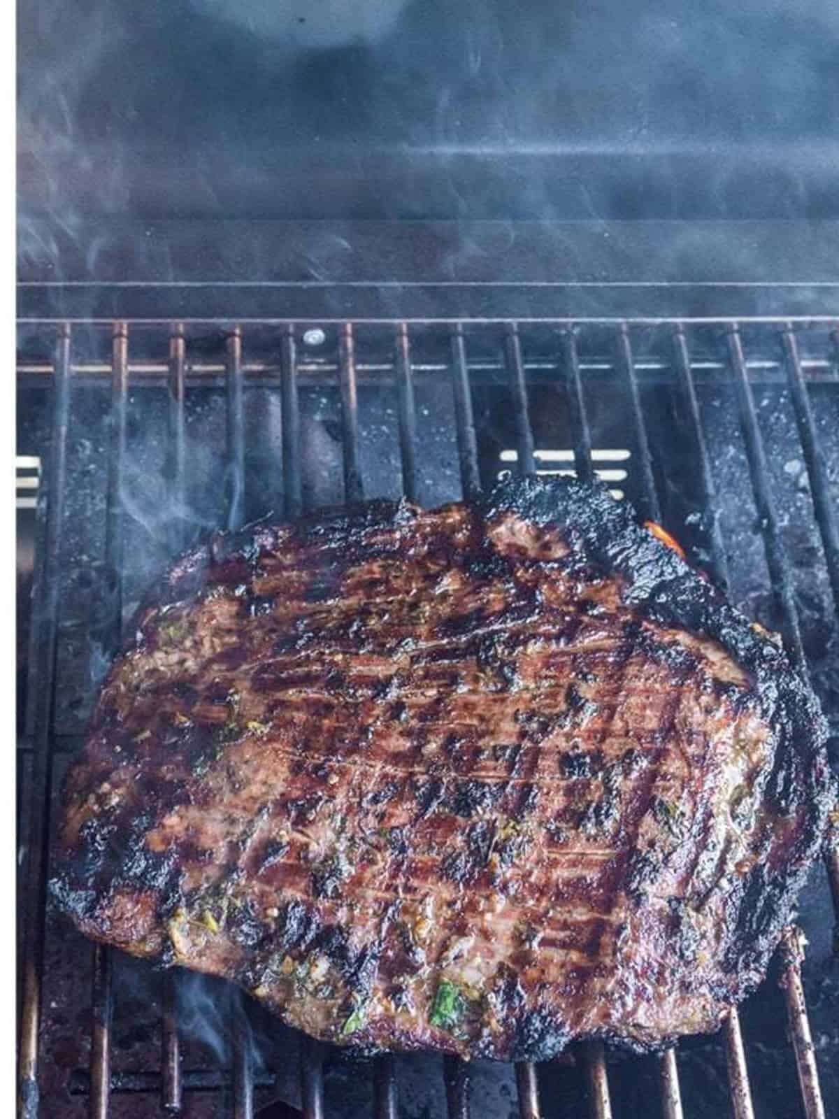 Marinated flank steak grilling over high heat with smoke rising.