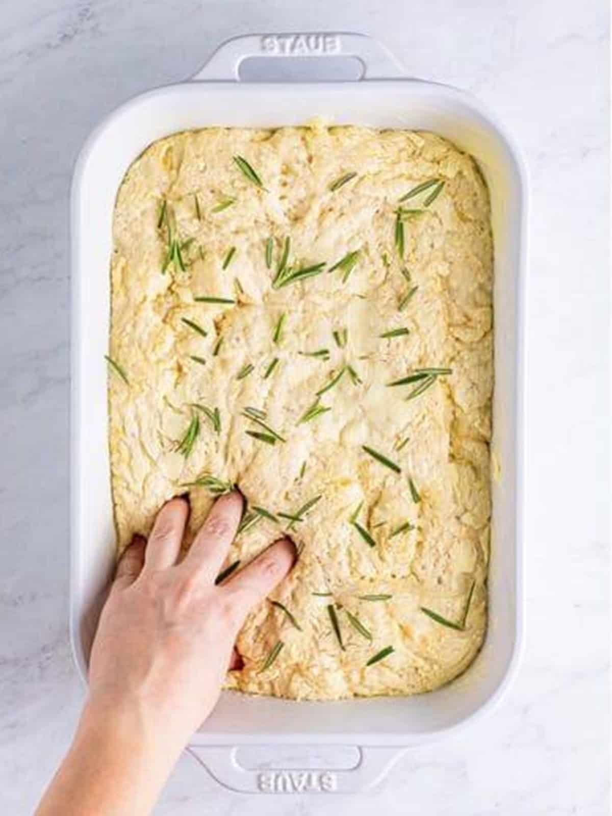 Dimpling the focaccia dough with fingers and scattering fresh rosemary on top.