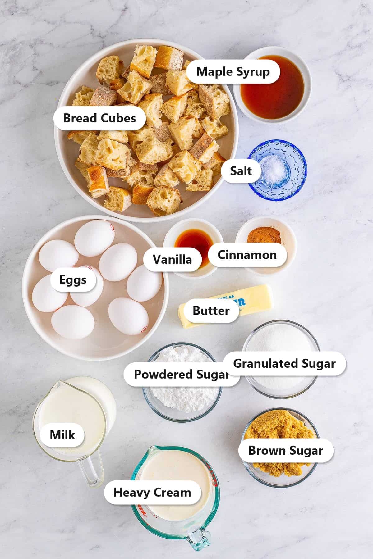 Labeled ingredients for overnight french toast casserole including french bread cubes, eggs, milk, heavy cream, sugars, cinnamon, and butter.