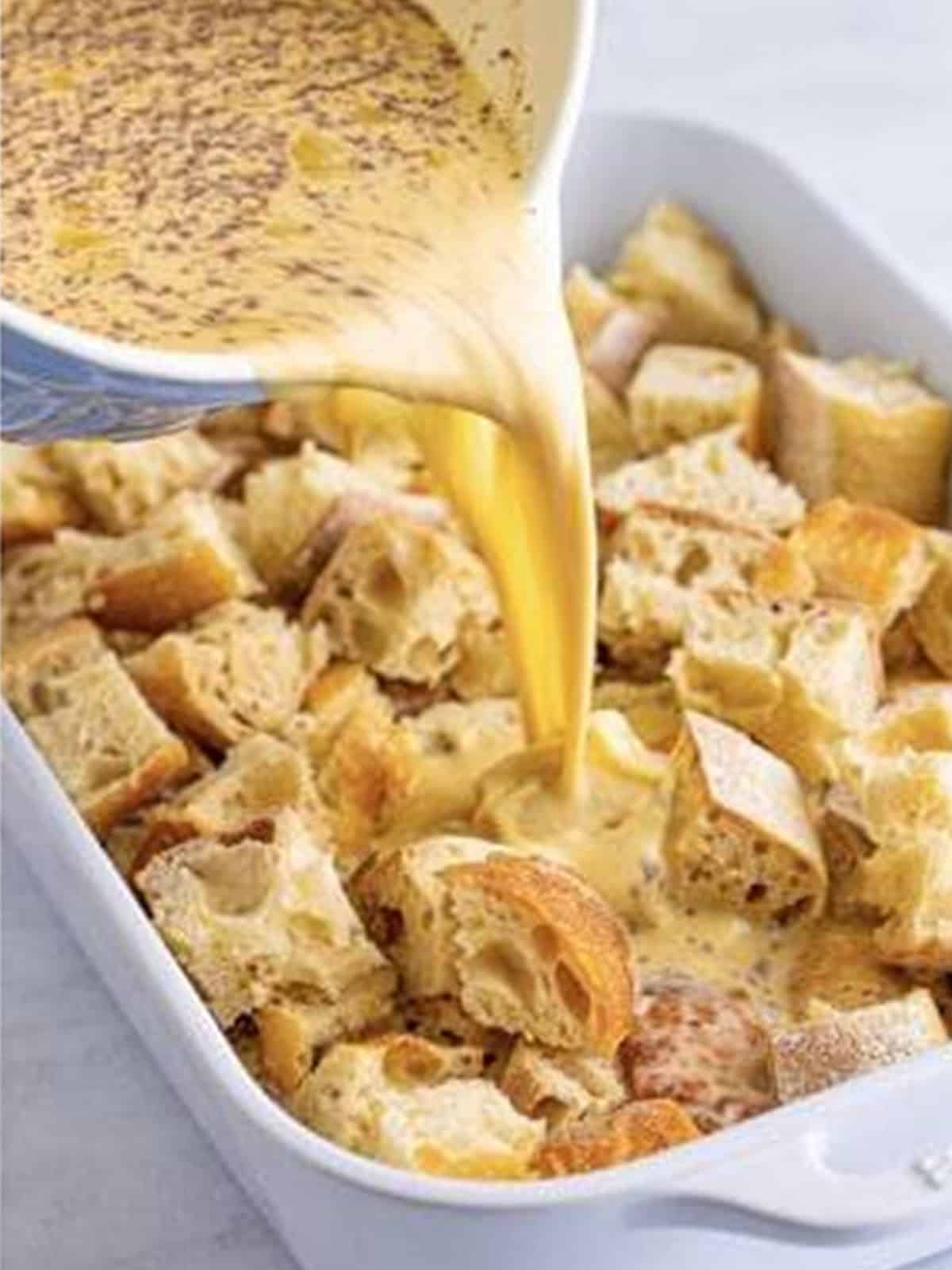 Custard being poured over cubed french bread in a baking dish.