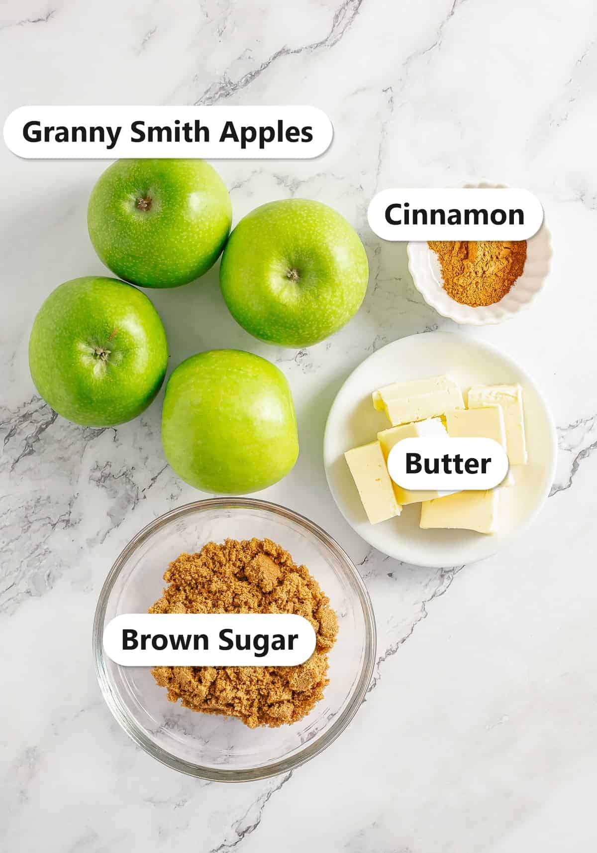 Ingredients to make fried apples labeled on a marble surface: granny smith apples, cinnamon, butter, and brown sugar.