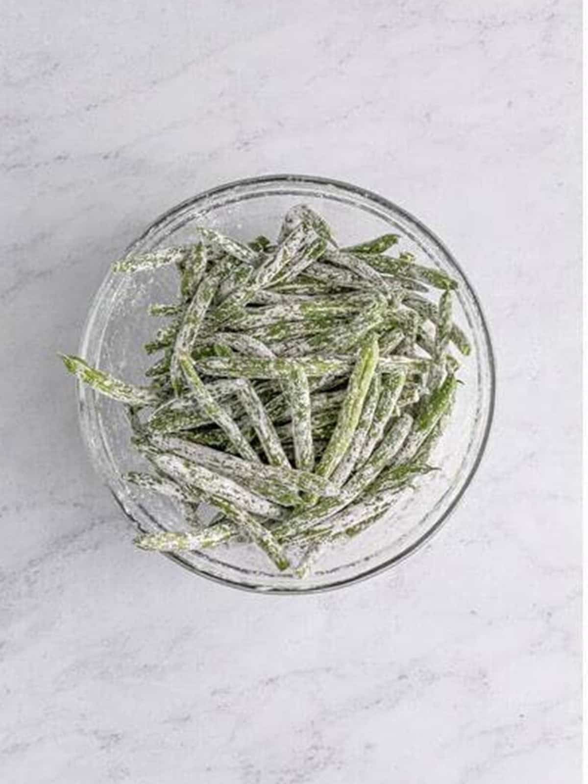 Fresh green beans tossed in cornstarch in a glass bowl.