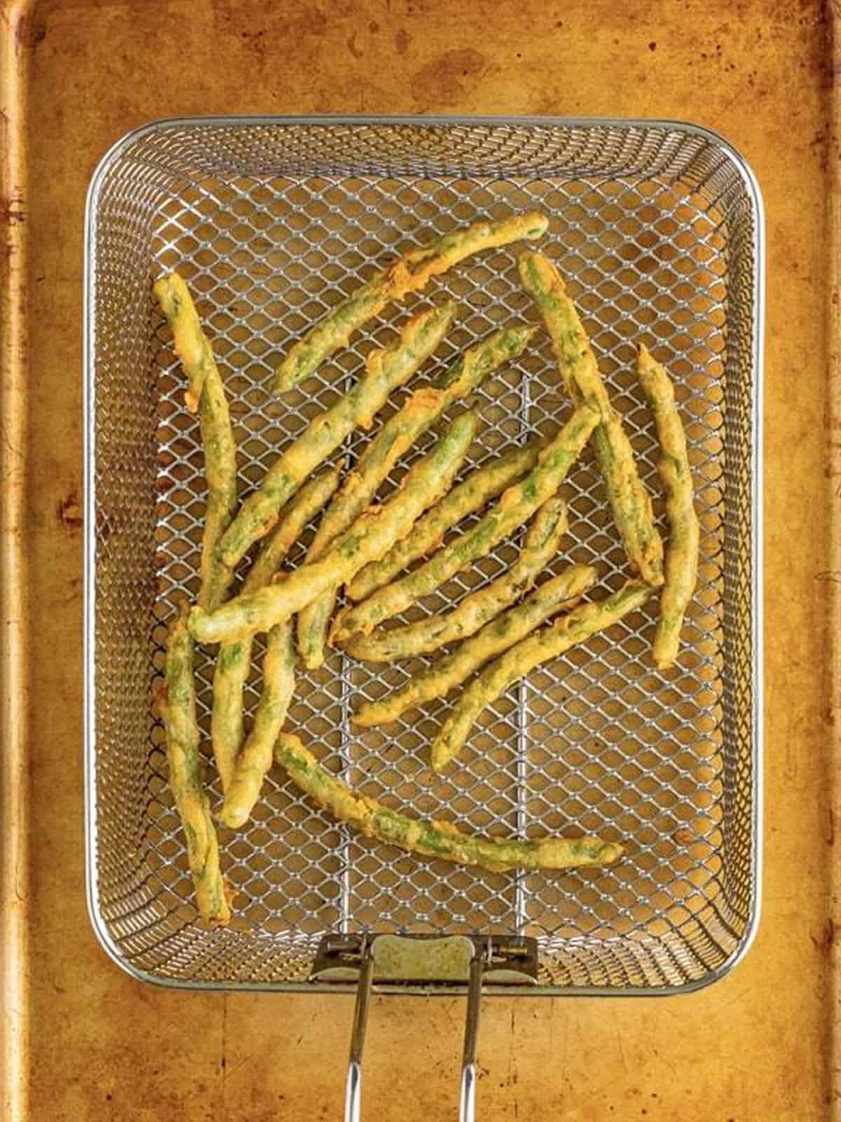 Battered green beans frying in a fryer basket until golden.