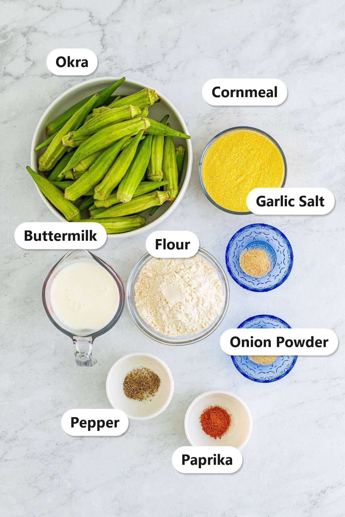 Labeled fried okra ingredients including fresh okra, cornmeal, flour, buttermilk, garlic salt, onion powder, pepper, and paprika on a marble counter.