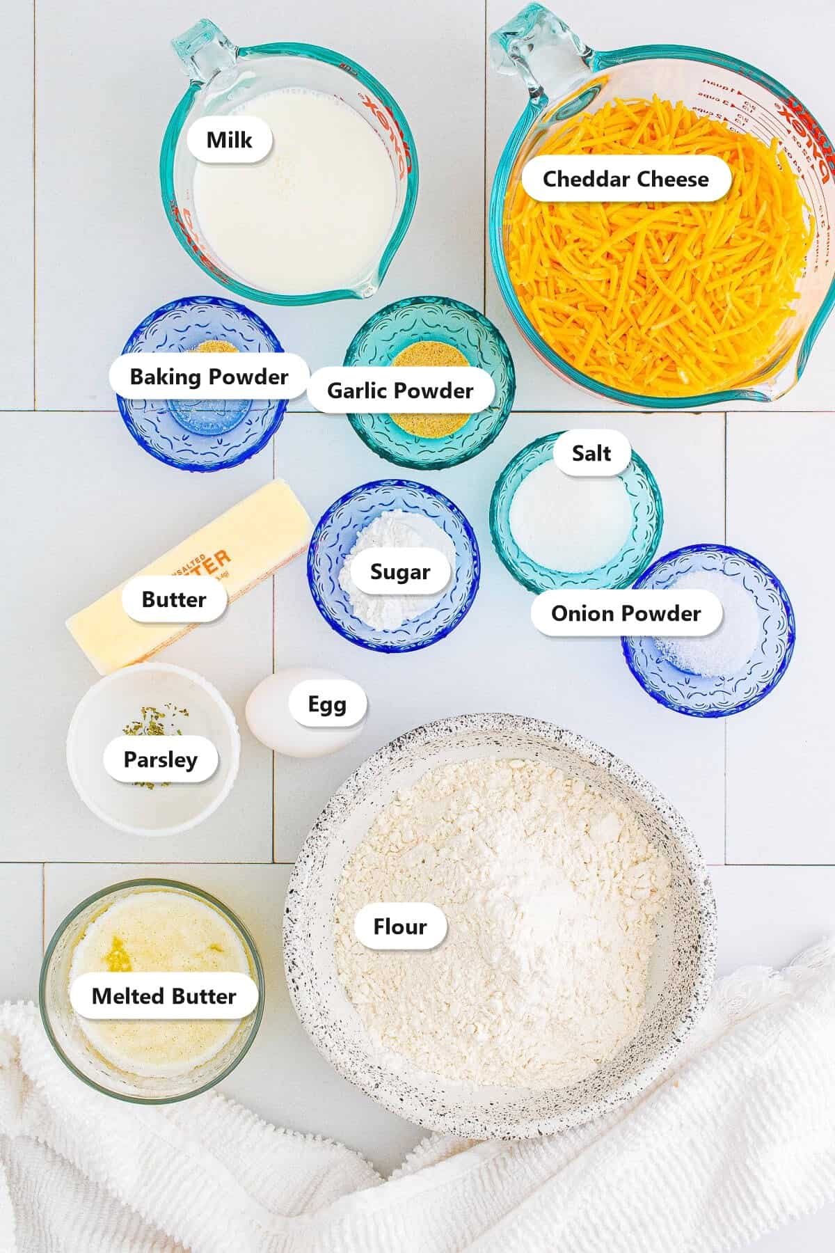 Labeled ingredients for garlic cheese bread including flour, shredded cheddar, milk, egg, butter, sugar, baking powder, salt, garlic powder, onion powder, and parsley.