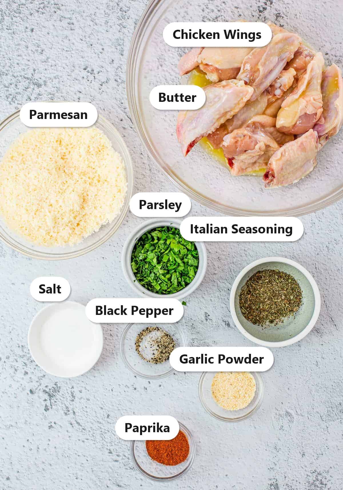 Ingredients to make garlic parmesan wings labeled on a marble surface including chicken wings, melted butter, parmesan, parsley, Italian seasoning, salt, black pepper, garlic powder, and paprika.