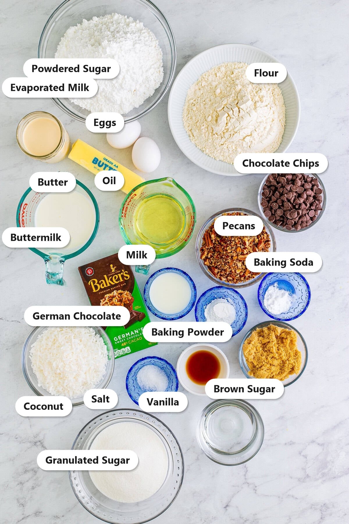German chocolate cake ingredients labeled on a marble counter including flour, sugars, German chocolate, coconut, and pecans.