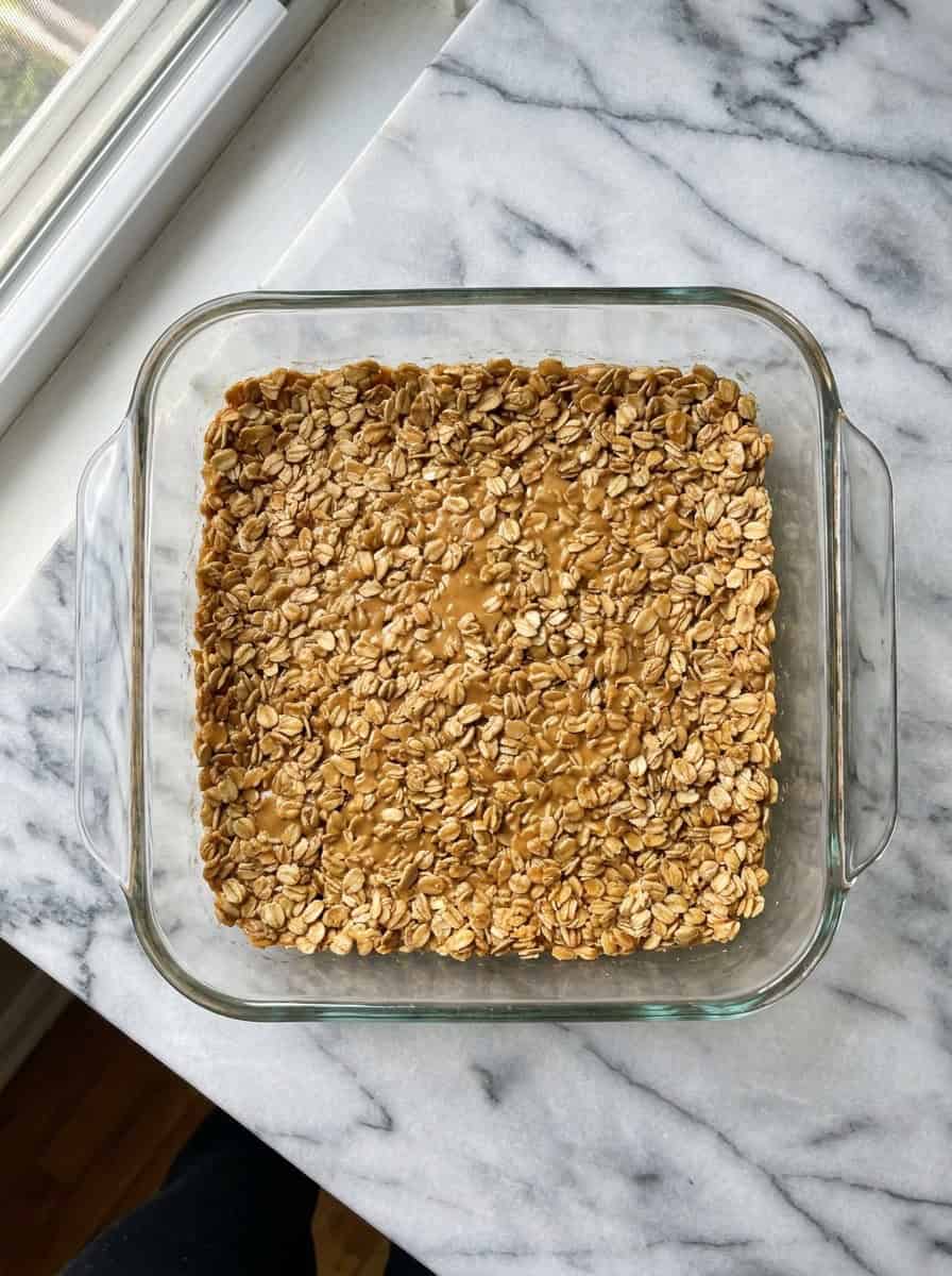 Granola mixture of oats, peanut butter, and honey pressed evenly into a square baking dish.