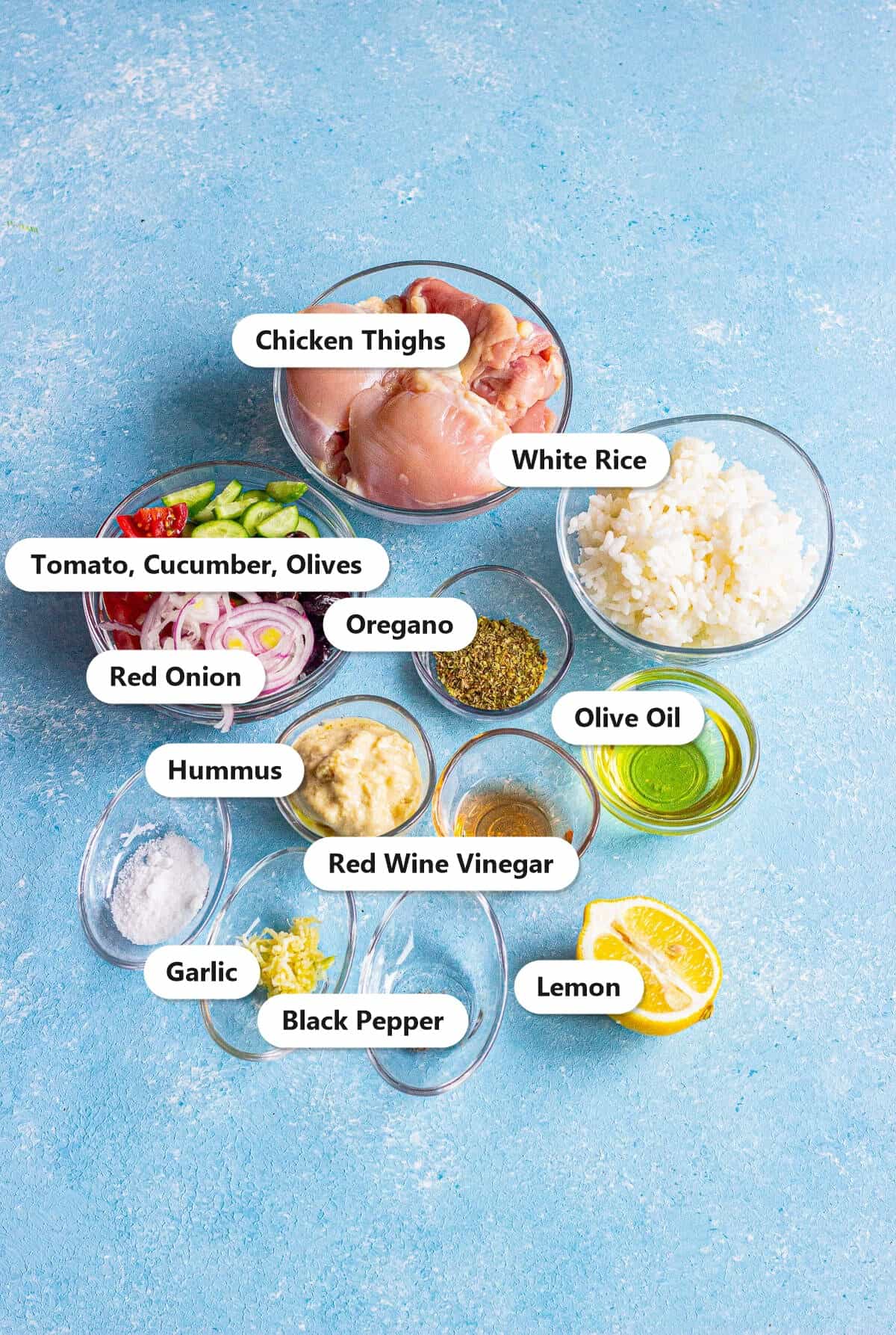 Labeled ingredients for a Greek chicken bowl including chicken thighs, rice, tomatoes, cucumber, olives, red onion, hummus, olive oil, lemon, and seasonings.