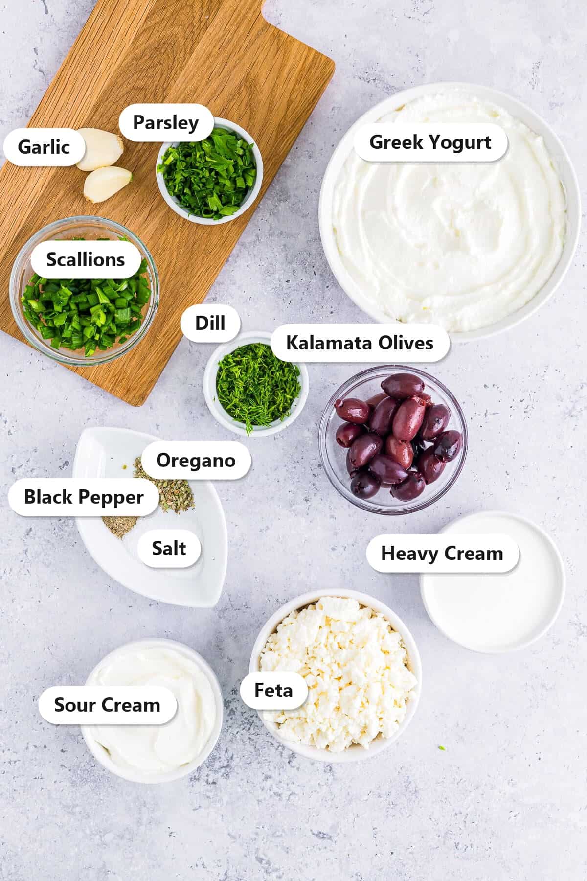 Ingredients to make greek yogurt dip labeled on a marble surface including greek yogurt, sour cream, heavy cream, feta, kalamata olives, garlic, dill, scallions, parsley, oregano, salt, and pepper.