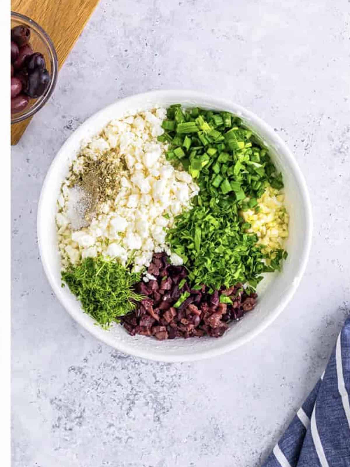 Greek yogurt with feta, kalamata olives, fresh herbs, garlic, and scallions added to a bowl.