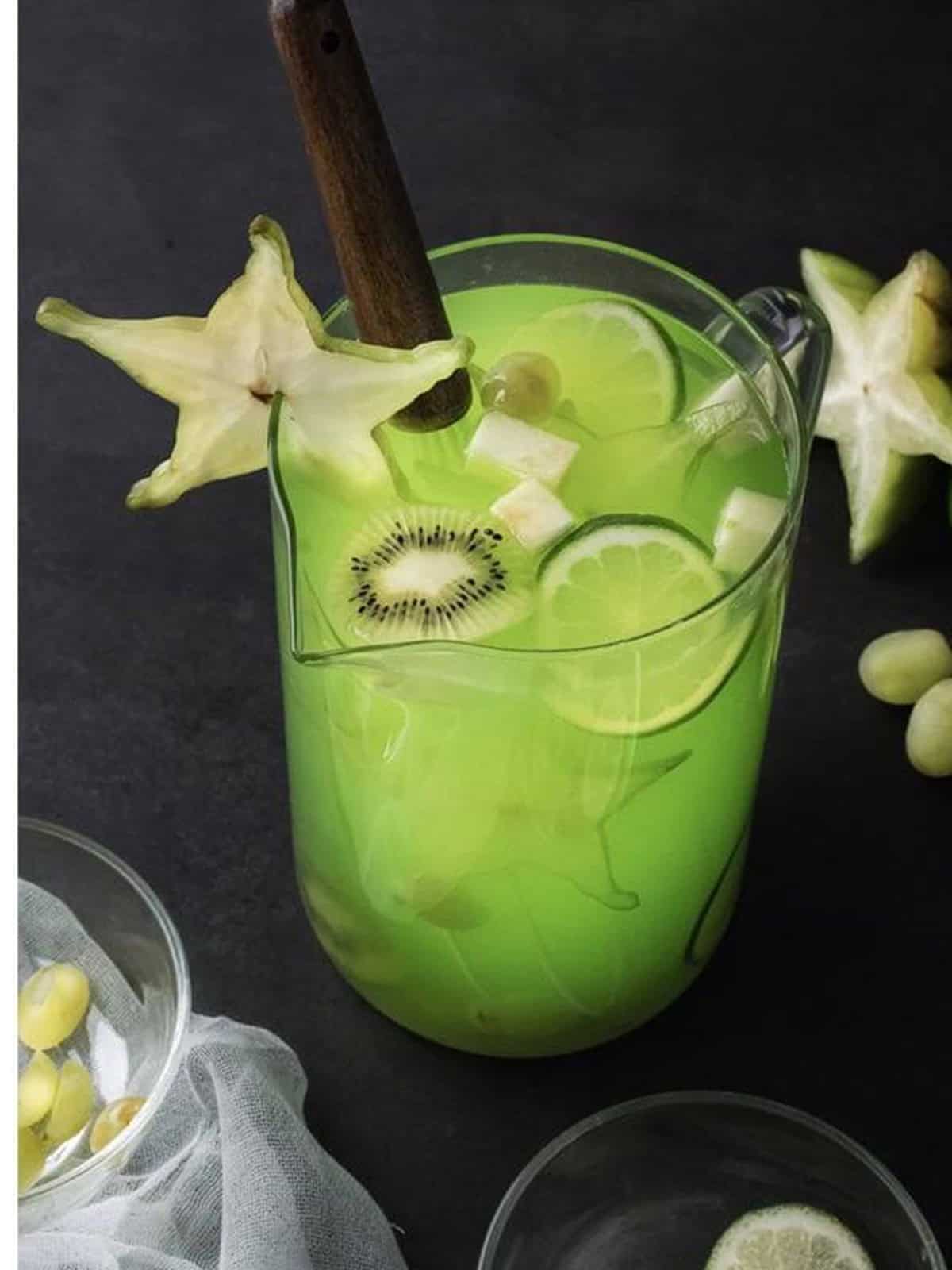 Finished green punch in a pitcher garnished with sliced kiwi, lime, and star fruit.