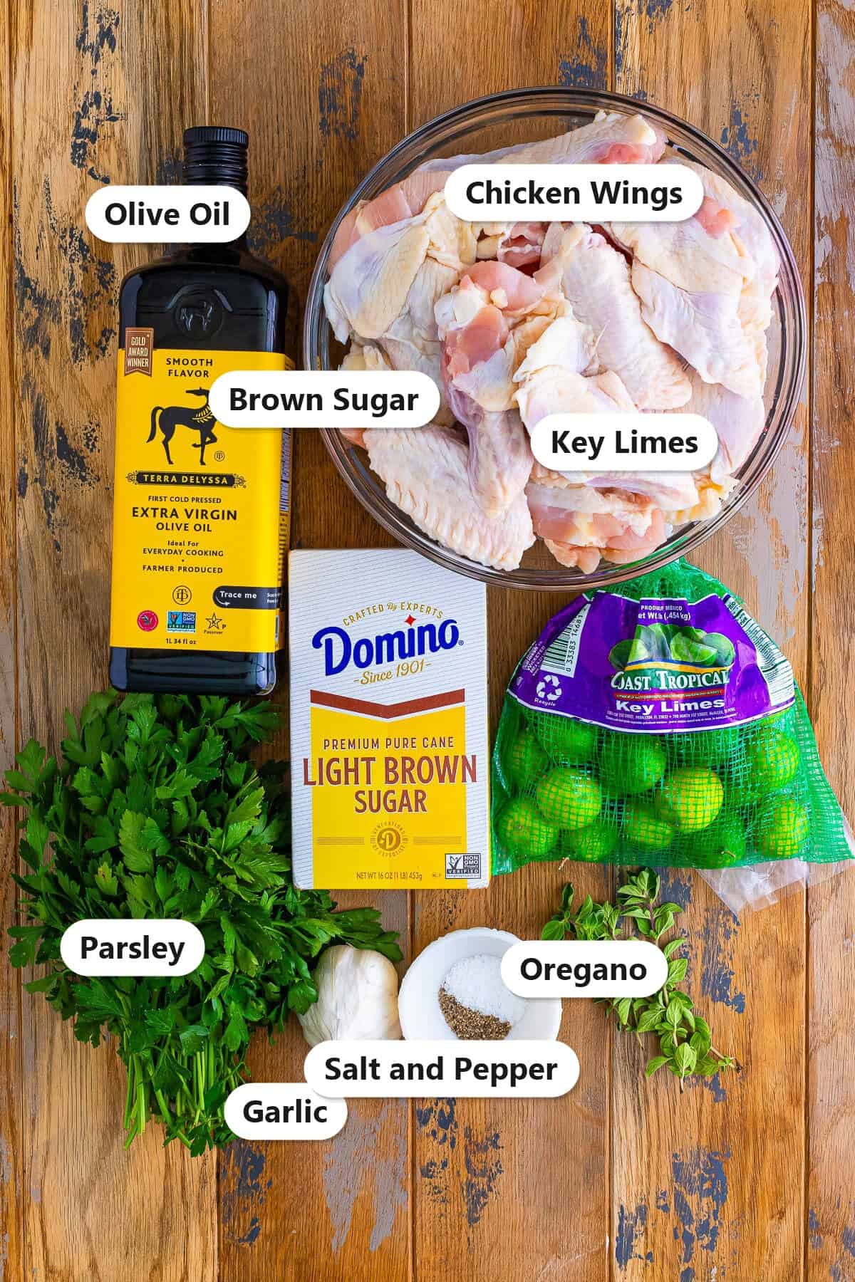 Grilled Chicken Wings ingredients including chicken wings, olive oil, brown sugar, key limes, garlic, parsley, oregano, salt, and pepper labeled on a board.