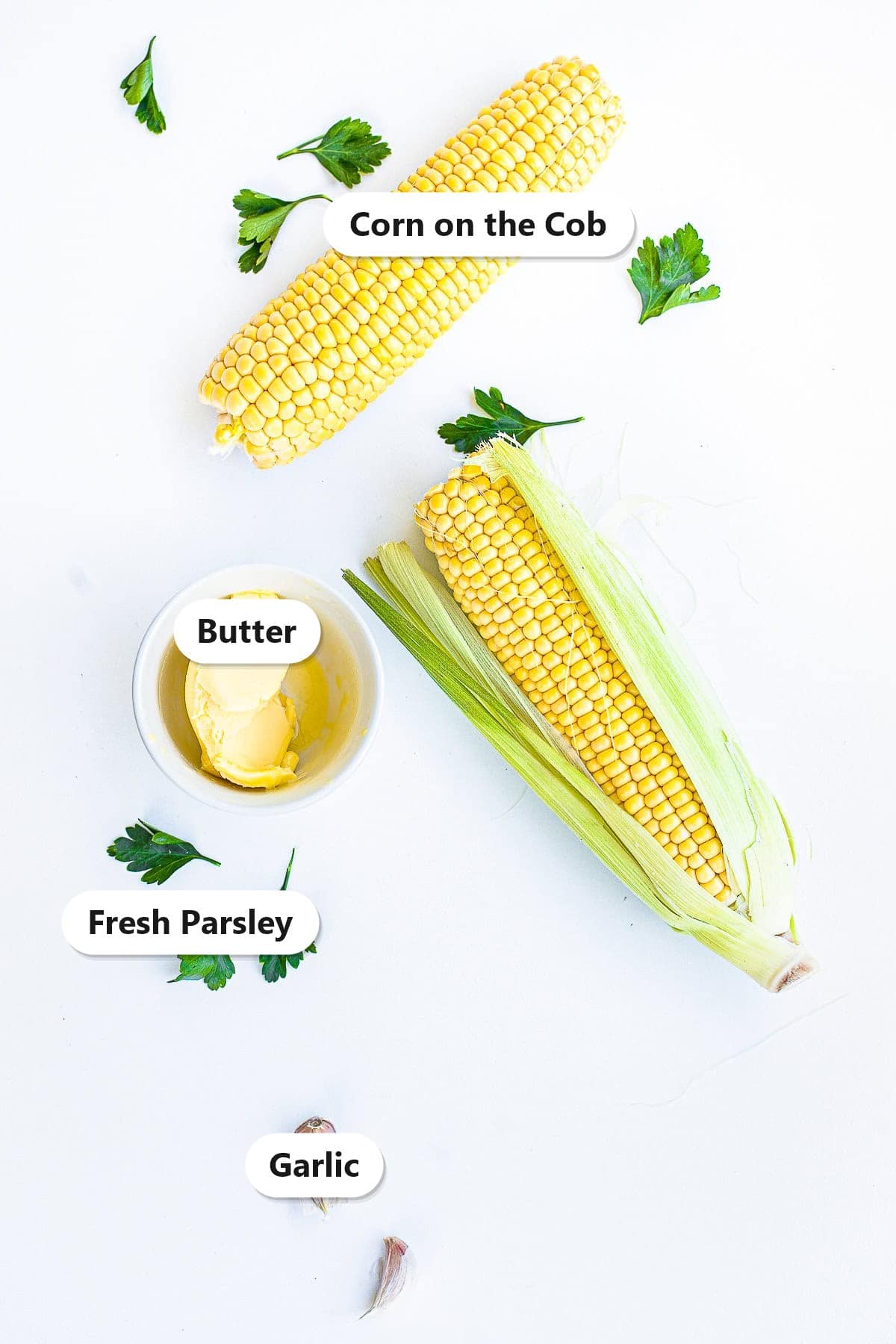 Labeled ingredients for grilled corn on the cob including fresh corn, butter, fresh parsley, and garlic on a white surface.