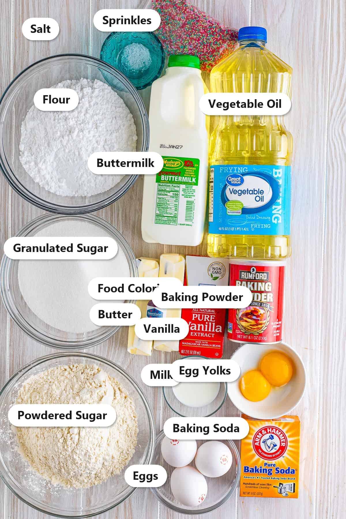 Labeled ingredients to make homemade birthday cupcakes including flour, granulated sugar, butter, vegetable oil, buttermilk, eggs, vanilla, powdered sugar, and sprinkles.