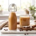 A glass bottle of homemade chocolate hazelnut coffee creamer beside a mug of coffee on a marble counter.