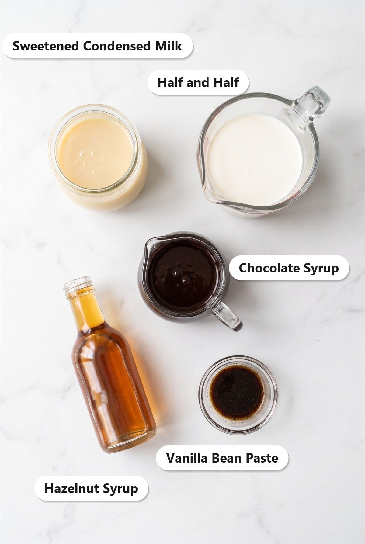 Labeled ingredients for homemade chocolate hazelnut coffee creamer including sweetened condensed milk, half and half, chocolate syrup, hazelnut syrup, and vanilla bean paste on a marble surface.