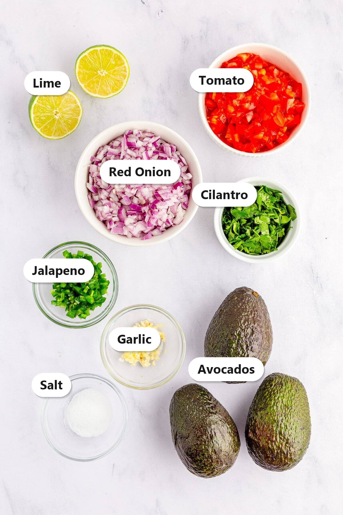 Guacamole ingredients including avocados, tomato, red onion, cilantro, jalapeno, garlic, lime, and salt labeled on a marble surface.