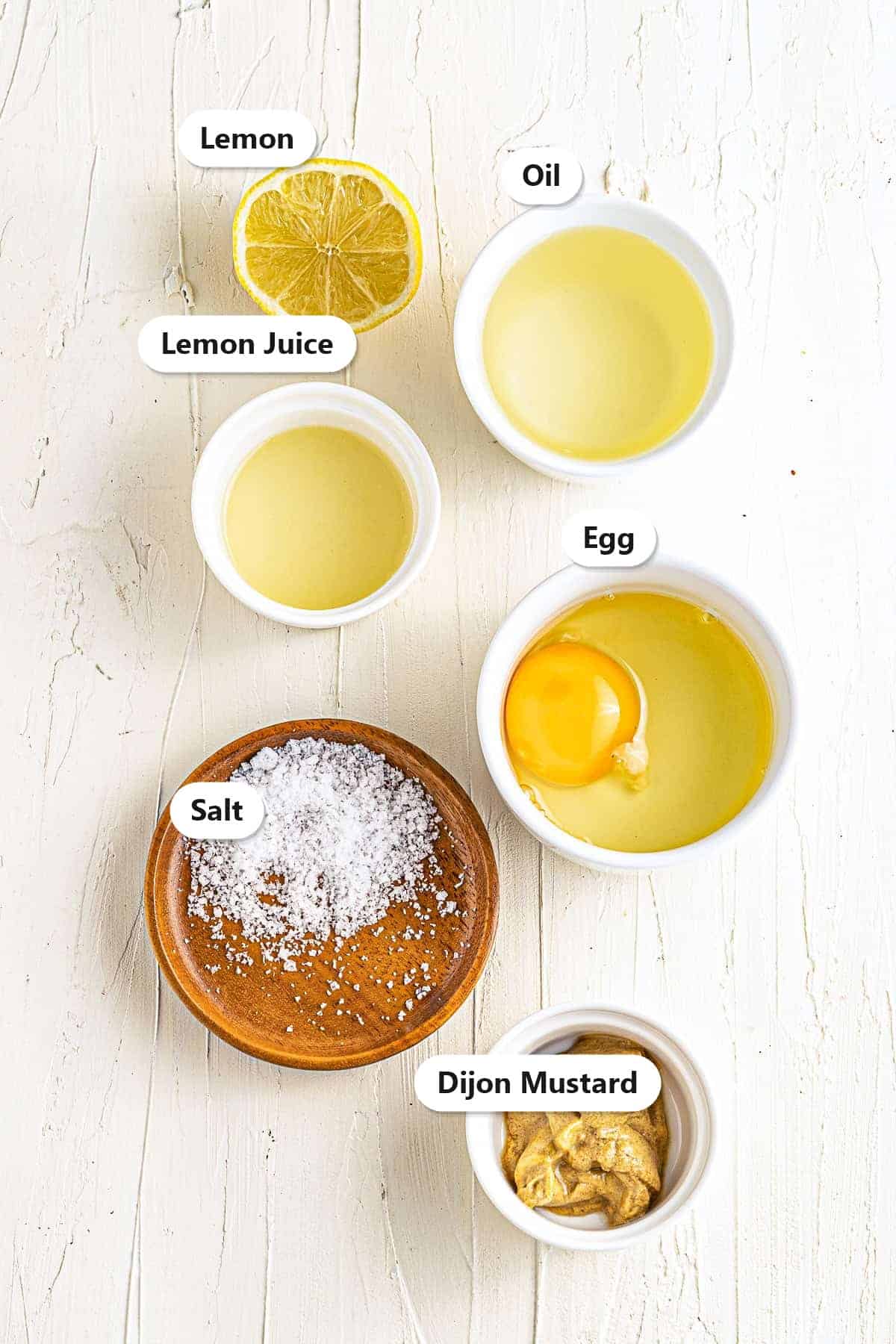 Homemade mayonnaise ingredients labeled on a white wood surface including a whole egg, canola oil, lemon juice, fresh lemon, Dijon mustard, and salt.