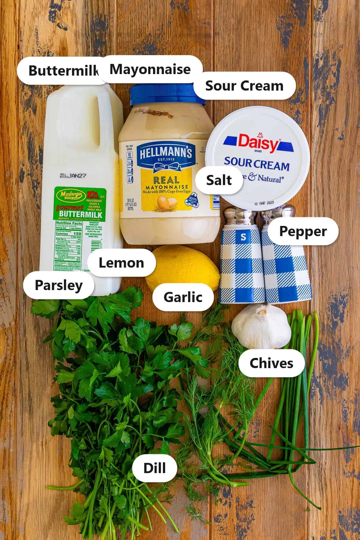 Labeled ingredients to make homemade ranch dressing including buttermilk, mayonnaise, sour cream, lemon, fresh dill, parsley, chives, garlic, salt, and pepper.