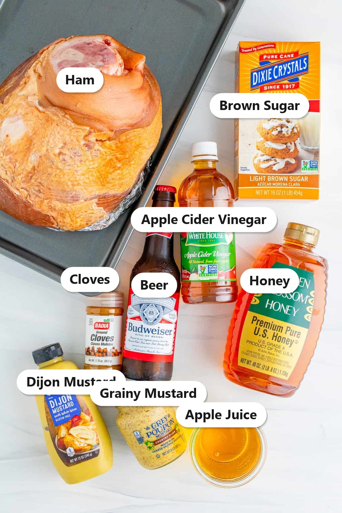 Labeled ingredients to make copycat honey baked ham including a smoked ham, beer, apple juice, honey, dijon and grainy mustard, brown sugar, apple cider vinegar, and cloves.