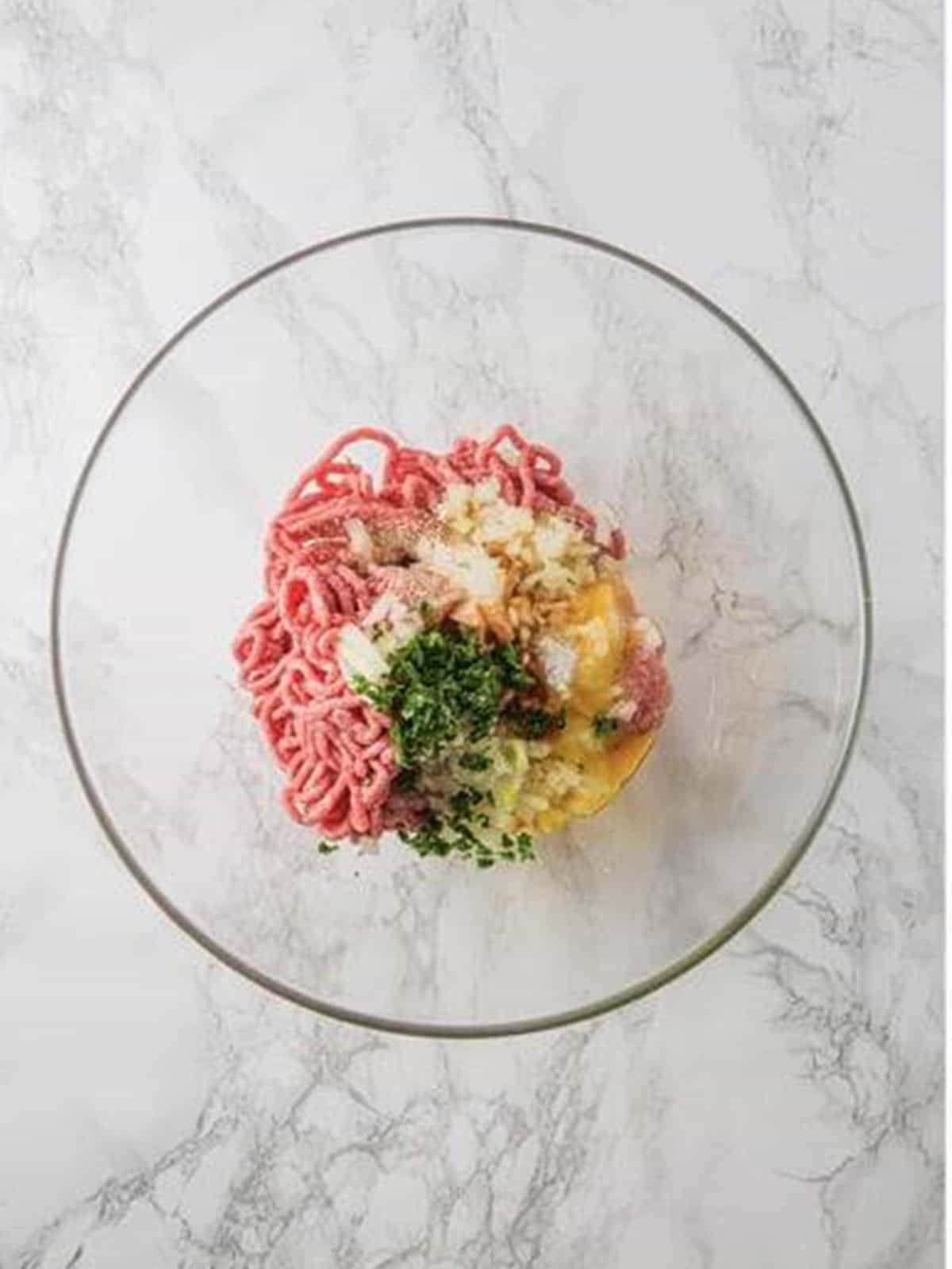Ground beef, egg, breadcrumbs, onion, and parsley combined in a glass bowl for meatballs.
