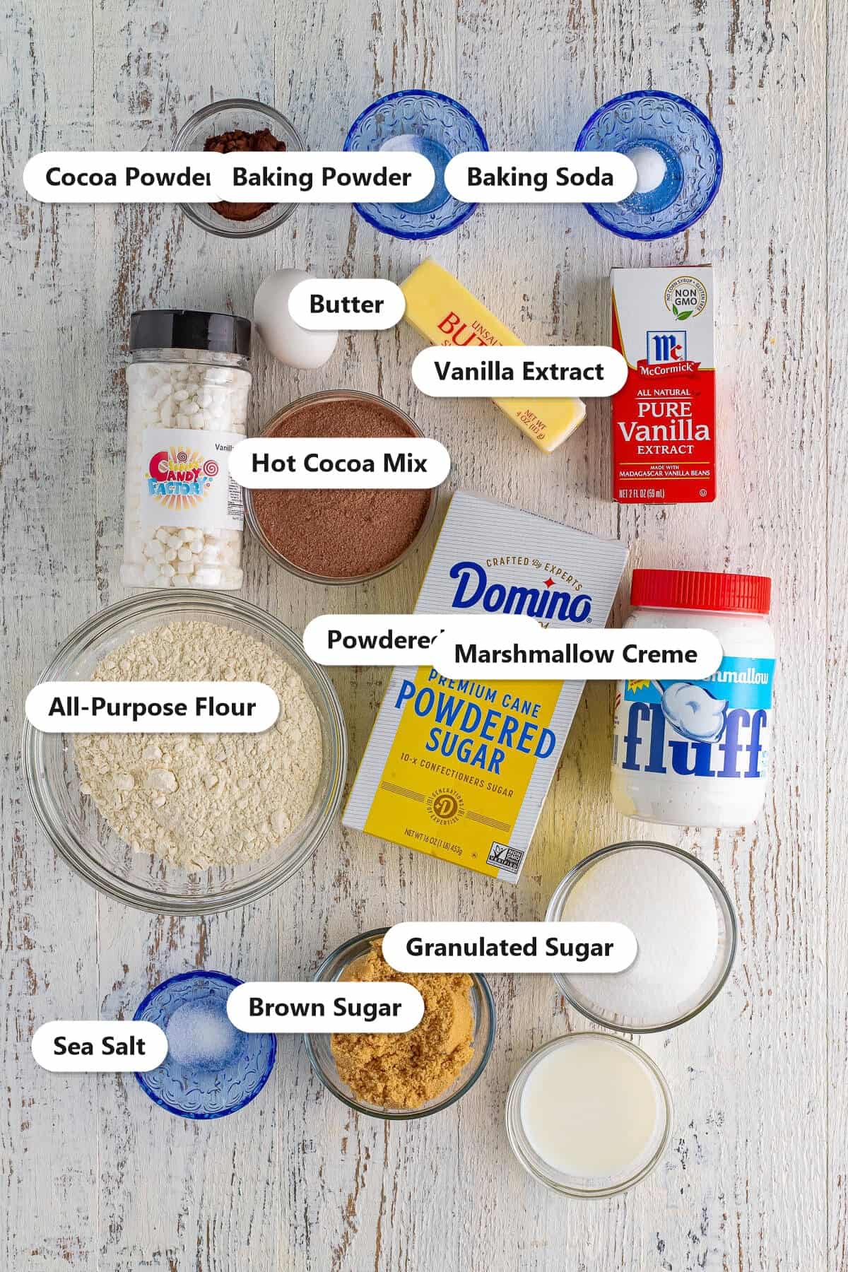 Labeled ingredients to make hot chocolate cookies: cocoa powder, baking powder, baking soda, butter, vanilla, hot cocoa mix, powdered sugar, marshmallow creme, flour, granulated sugar, brown sugar, and sea salt.