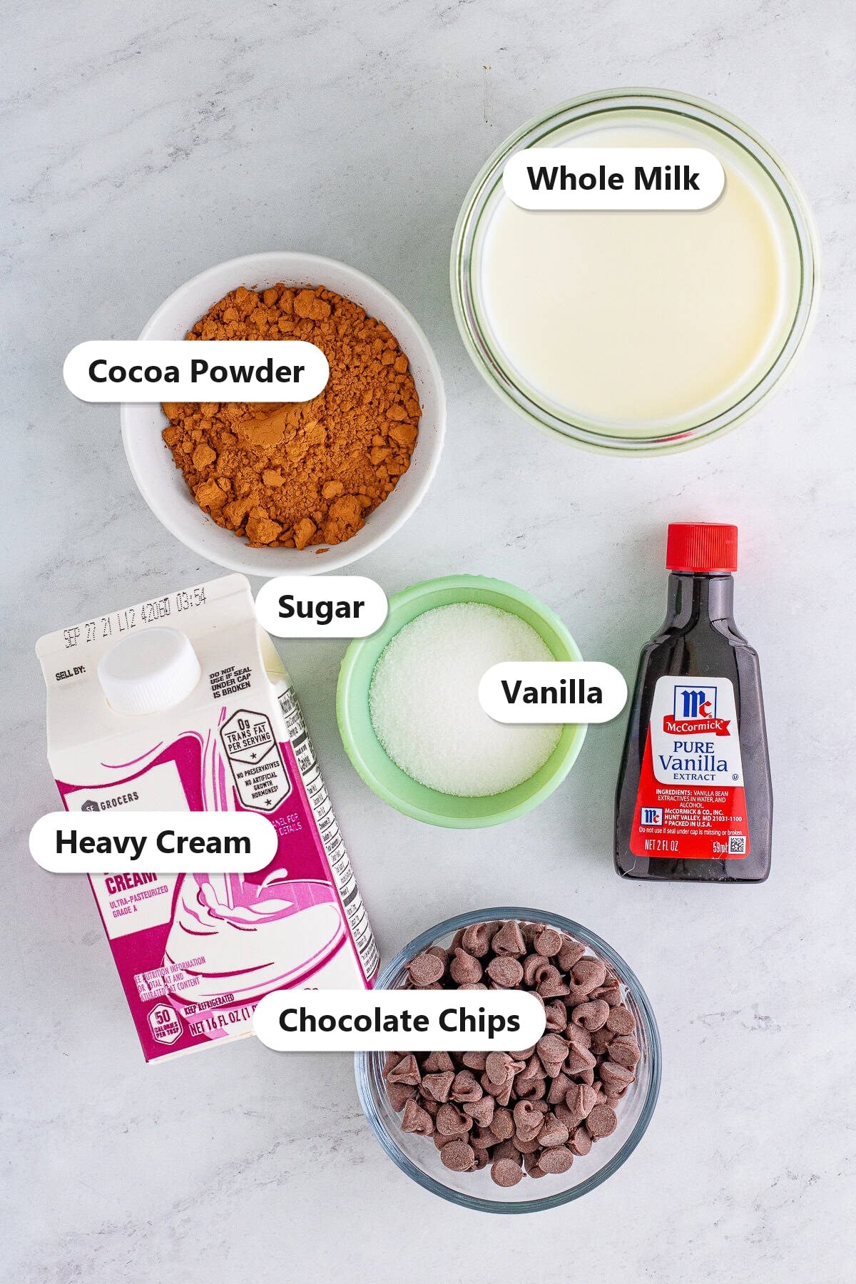 Hot chocolate ingredients including whole milk, heavy cream, cocoa powder, sugar, milk chocolate chips, and vanilla labeled on a marble surface.