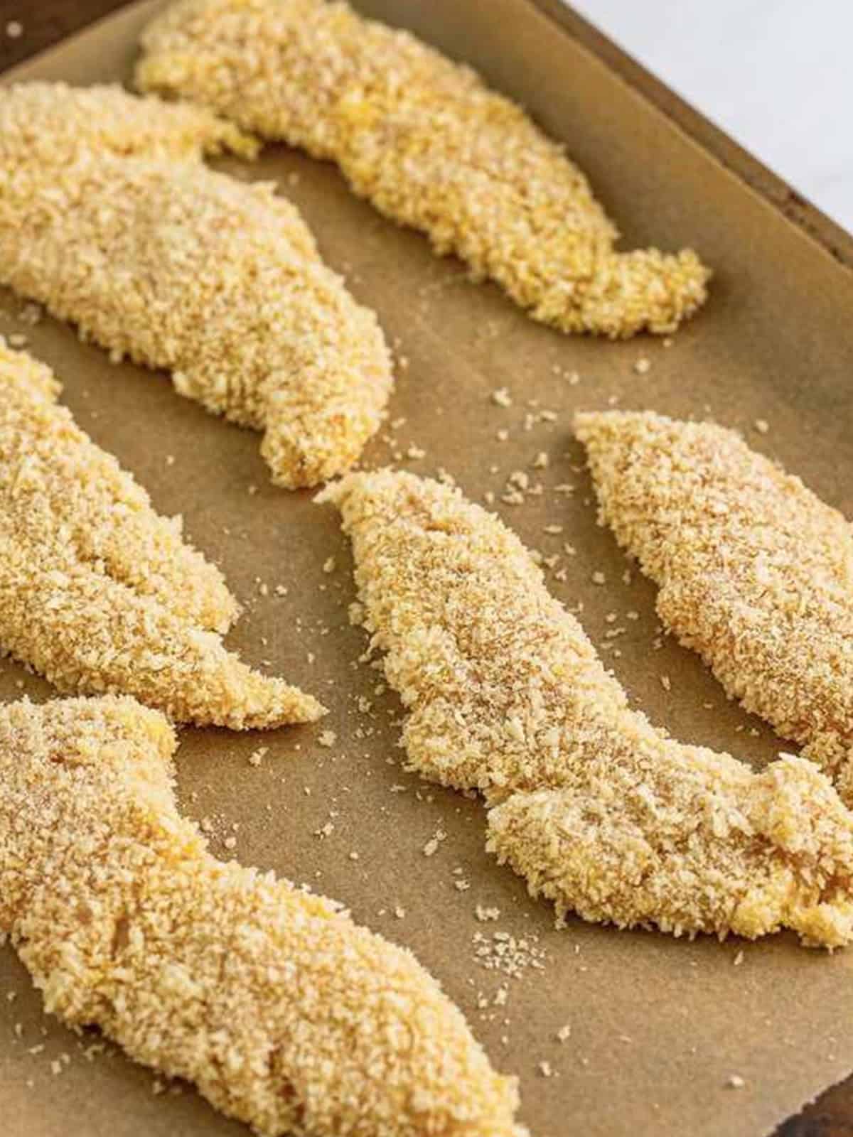 Breaded chicken tenders lined up on a parchment-lined sheet tray for Hot Honey Chicken.
