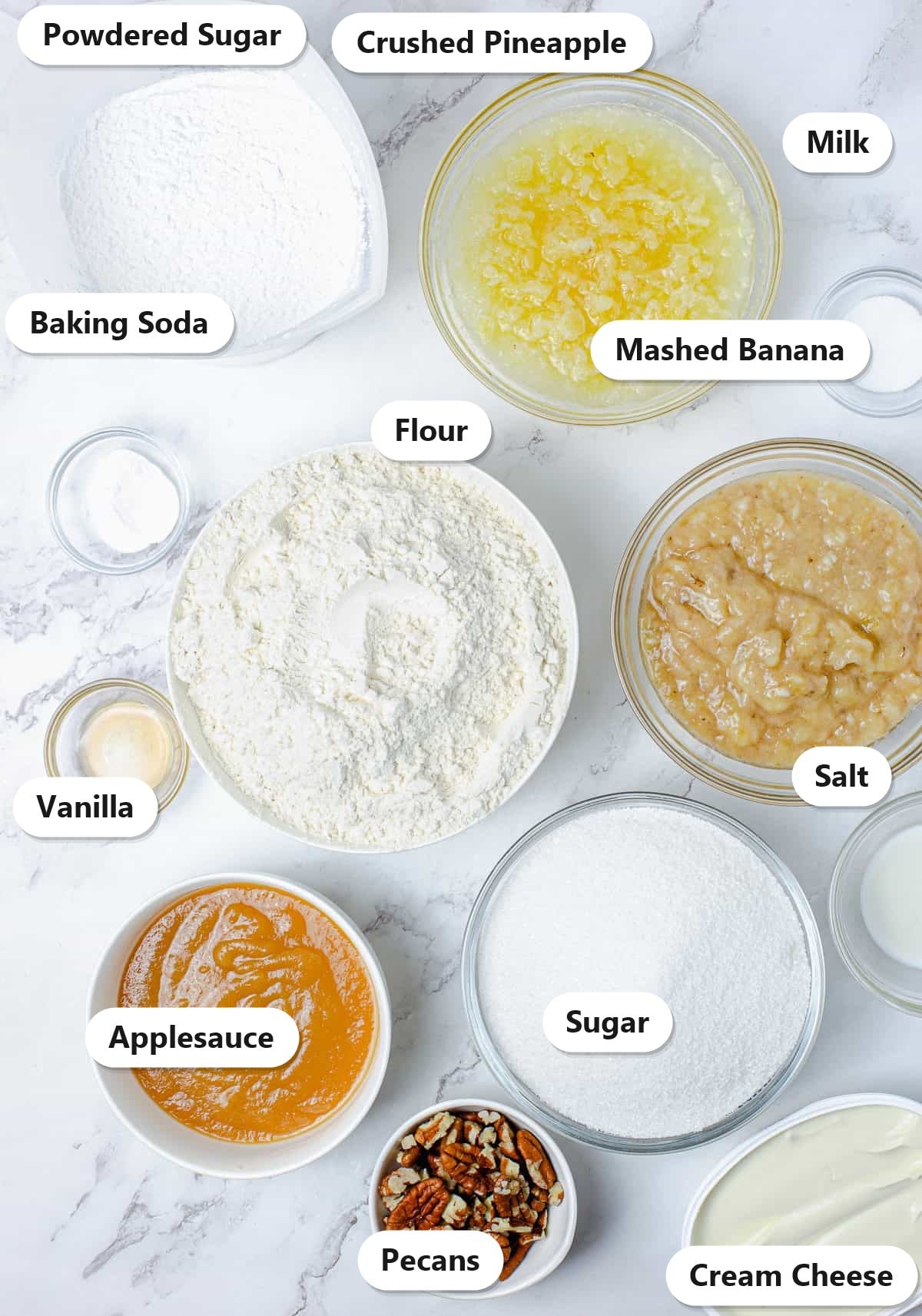 Labeled hummingbird cake ingredients including flour, sugar, mashed banana, crushed pineapple, applesauce, pecans, and cream cheese on a marble counter.