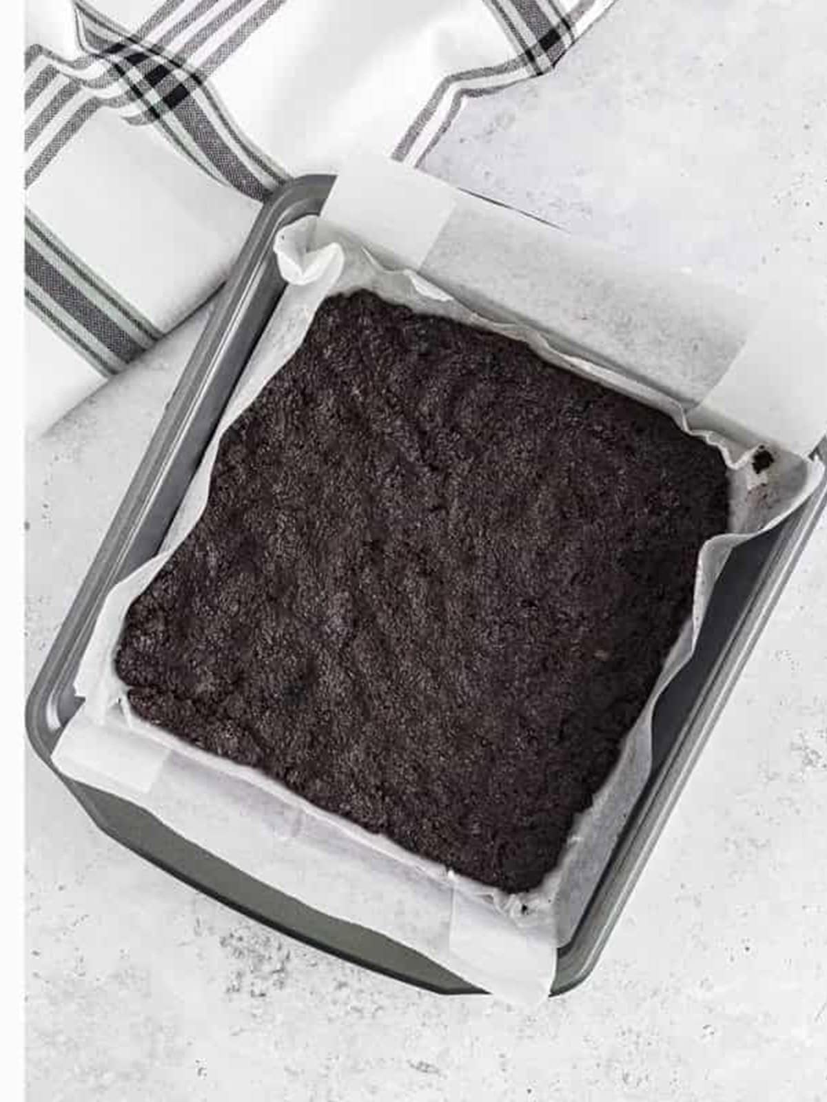 Oreo crust pressed into a parchment lined square pan for frozen dirt cake.