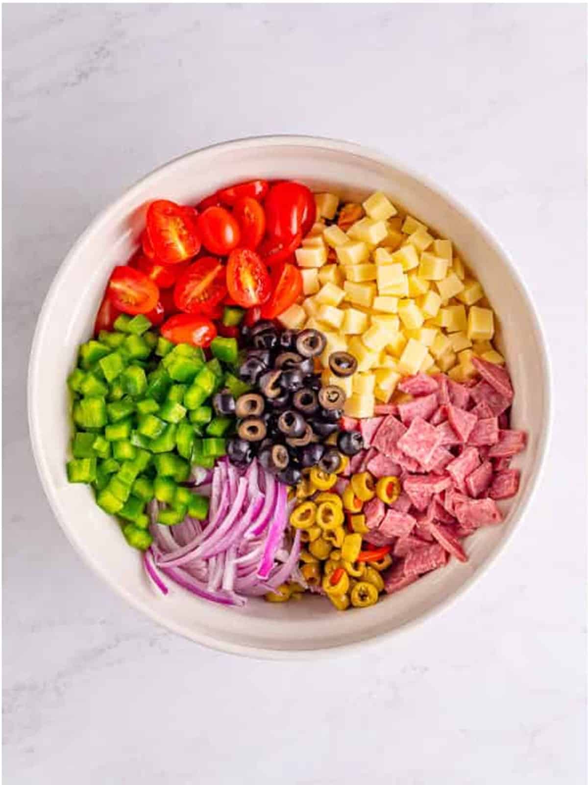 Grape tomatoes, mozzarella, green pepper, red onion, salami, and olives combined in a bowl for Italian pasta salad.