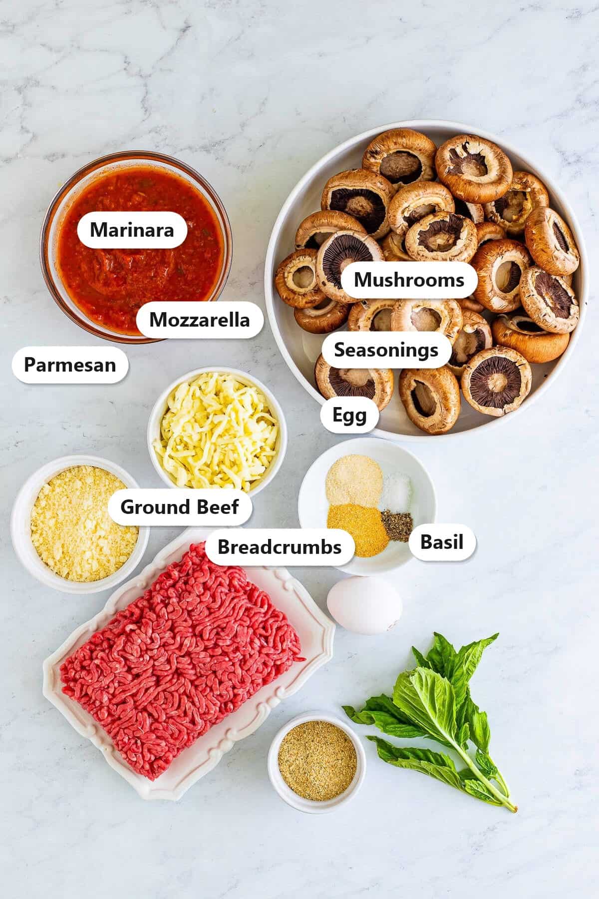 Labeled ingredients for Italian stuffed mushrooms including baby bella mushrooms, ground beef, marinara, parmesan, mozzarella, breadcrumbs, egg, seasonings, and basil.