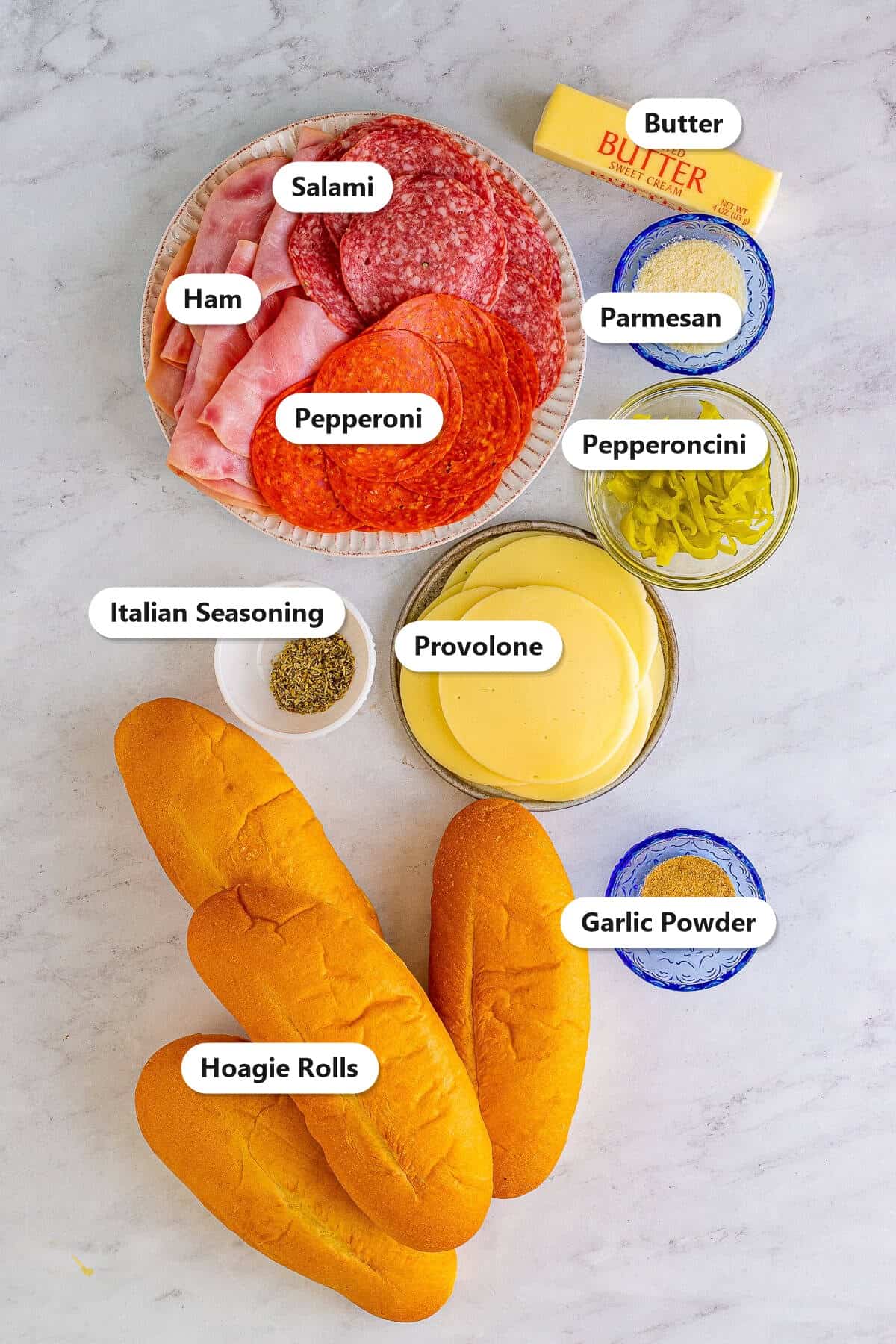Italian sub ingredients labeled on marble including hoagie rolls, salami, ham, pepperoni, provolone, butter, parmesan, garlic powder, Italian seasoning, and pepperoncini.