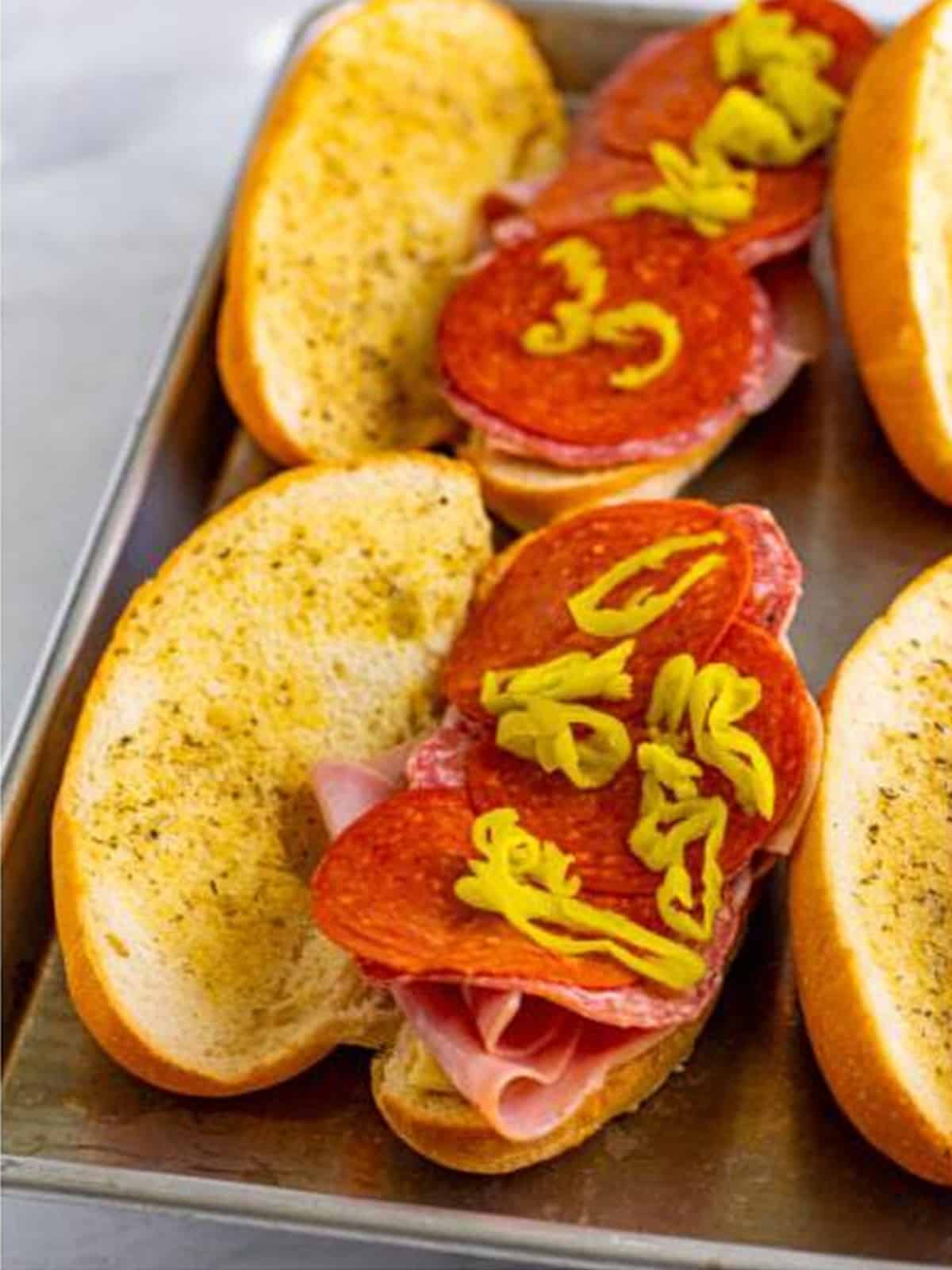 Layering ham, salami, pepperoni, and pepperoncini onto the sub rolls.