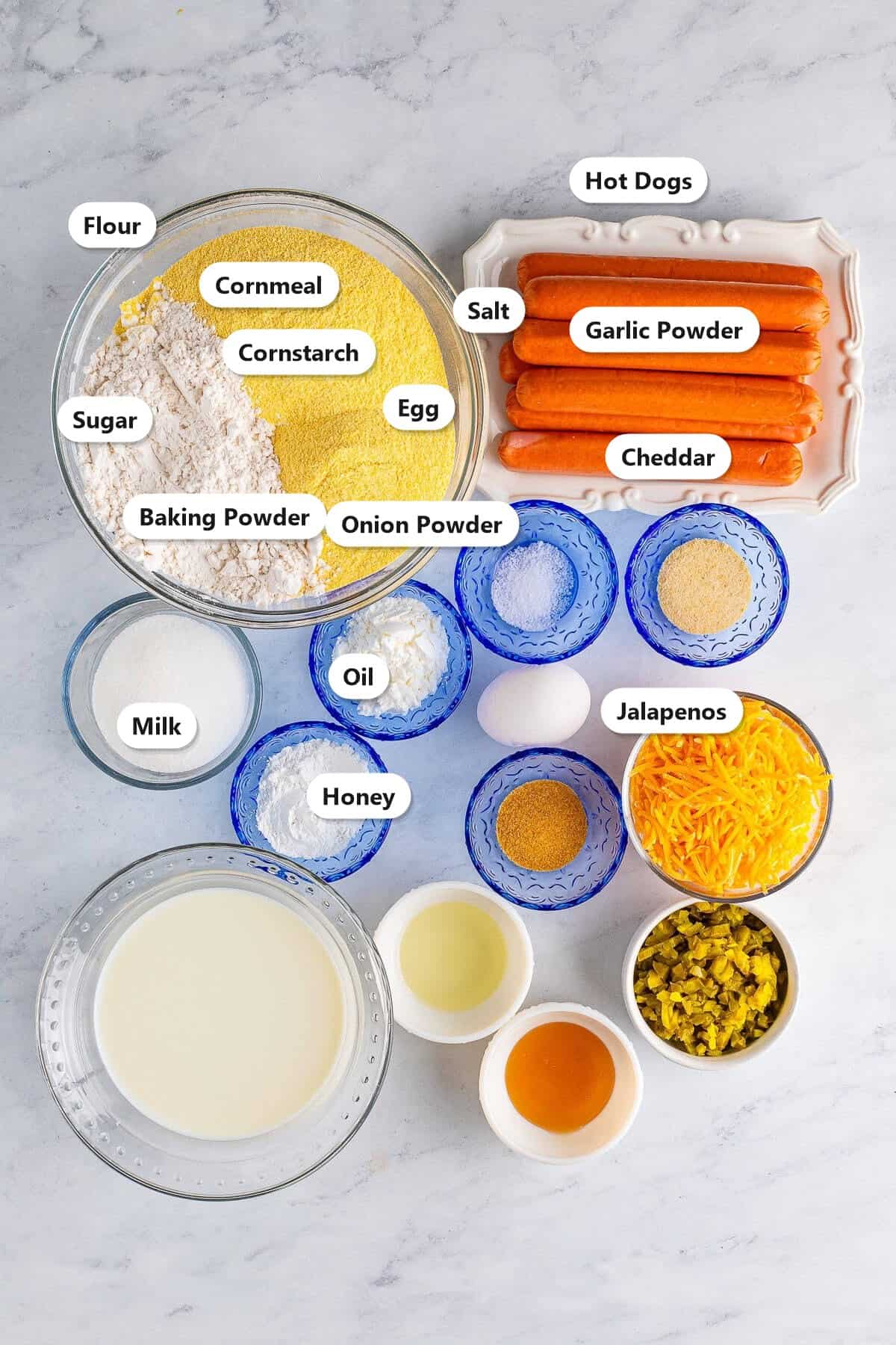 Labeled ingredients for homemade jalapeno cheddar corn dogs including cornmeal, flour, hot dogs, cheddar, jalapenos, milk, egg, oil, honey, sugar, and seasonings.