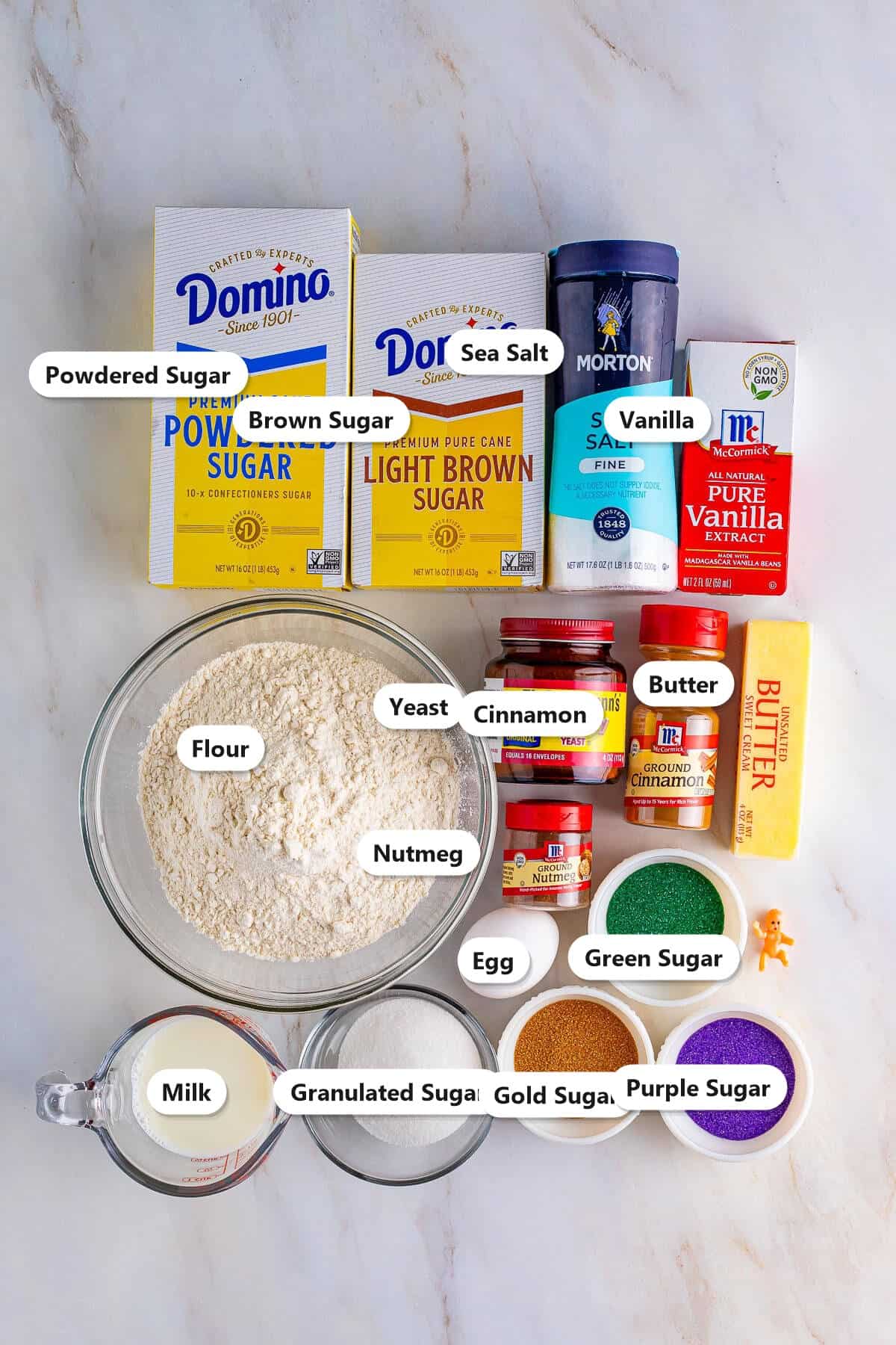 King cake ingredients labeled on marble including flour, yeast, butter, milk, eggs, sugar, cinnamon, nutmeg, vanilla, and purple, green, and gold sanding sugars.
