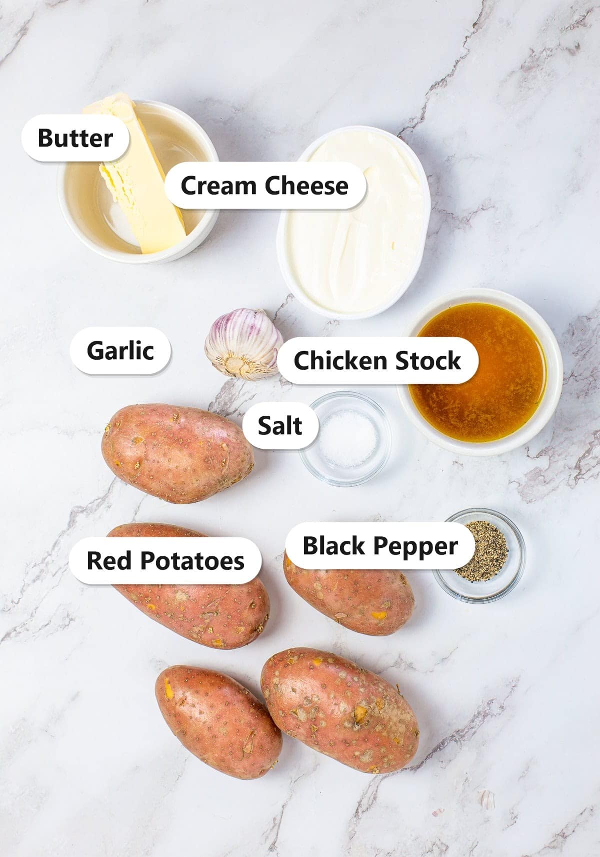 Labeled ingredients to make creamy mashed potatoes including red potatoes, butter, cream cheese, garlic, chicken stock, salt, and black pepper.